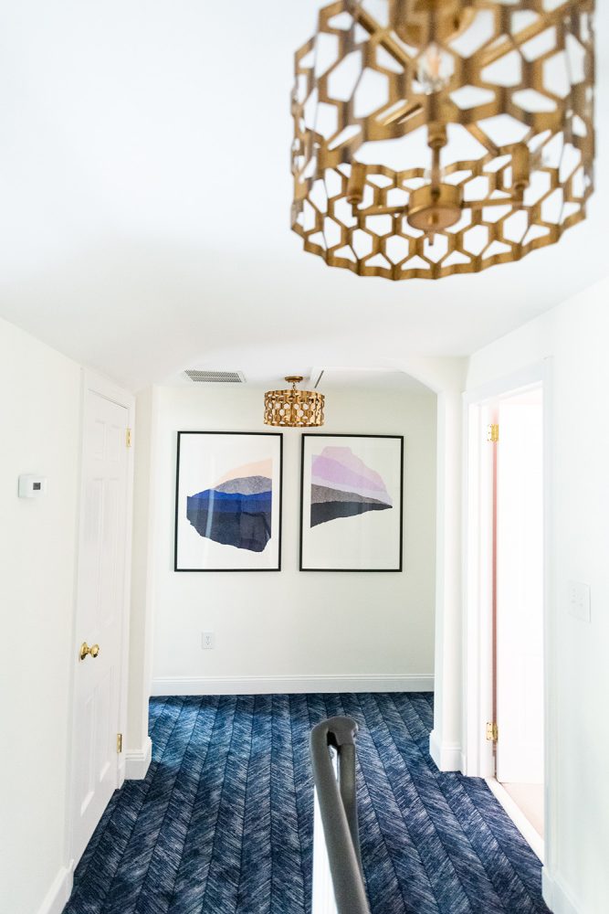 Two pieces of abstract art at the end of a white-walled hallway above blue carpet. Piece on left has beige, grey and blue shapes, piece on the right has purples, gray and black. Brass light fixtures hang from ceiling.
