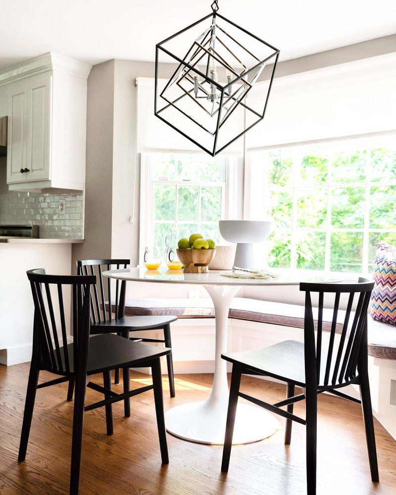 Kitchen banquette built beneath a bay window. The banquette curves to match the angled placement of the windowpanes, and is upholstered in a tweedy mauve fabric. White tulip table with black wooden chairs in front and pendant lamp is several inset angular cubes in dark metal.