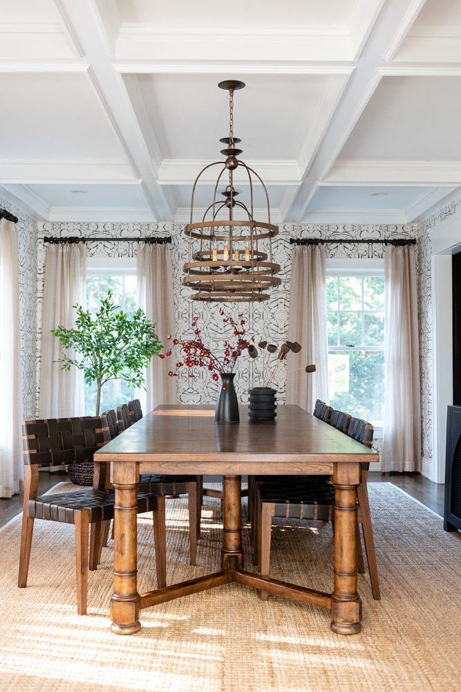 Dining room with rectangular wooden dining table and wooden chairs with woven black leather seats. Area rug of braided natural fibers on floor. Walls have white wallpaper with black scribbled geometric design. Birdcage-like brown metal chandelier above table.