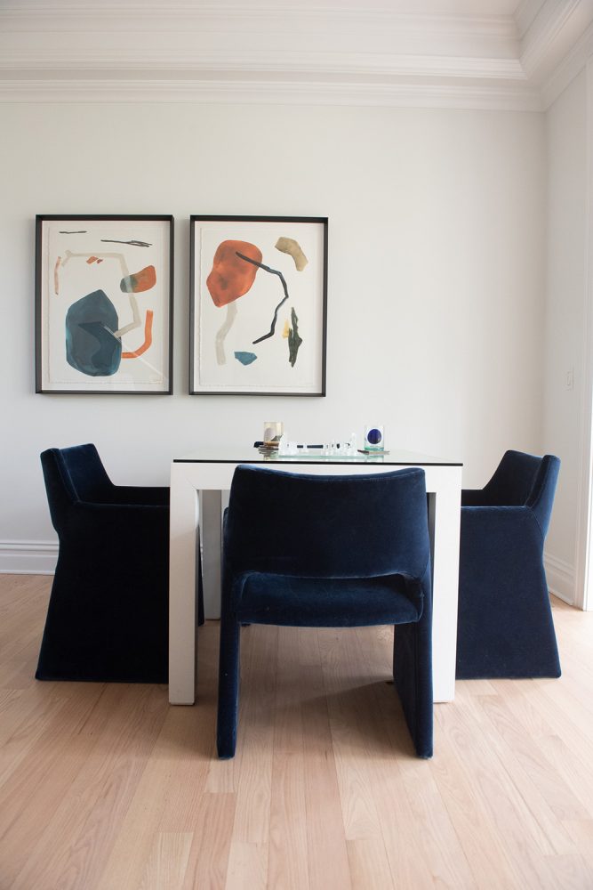 Two pieces of abstract art with blue, orange, and gray figures above a white table with blue upholstered chairs.
