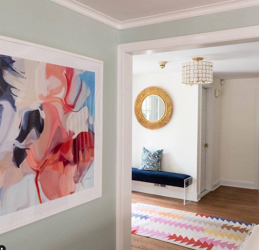 Photo of an entry hall with abstract pastel art hung on one wall, a pastel geometric patterned runner on the floor, a blue upholstered bench and a round textured brass mirror on another wall.