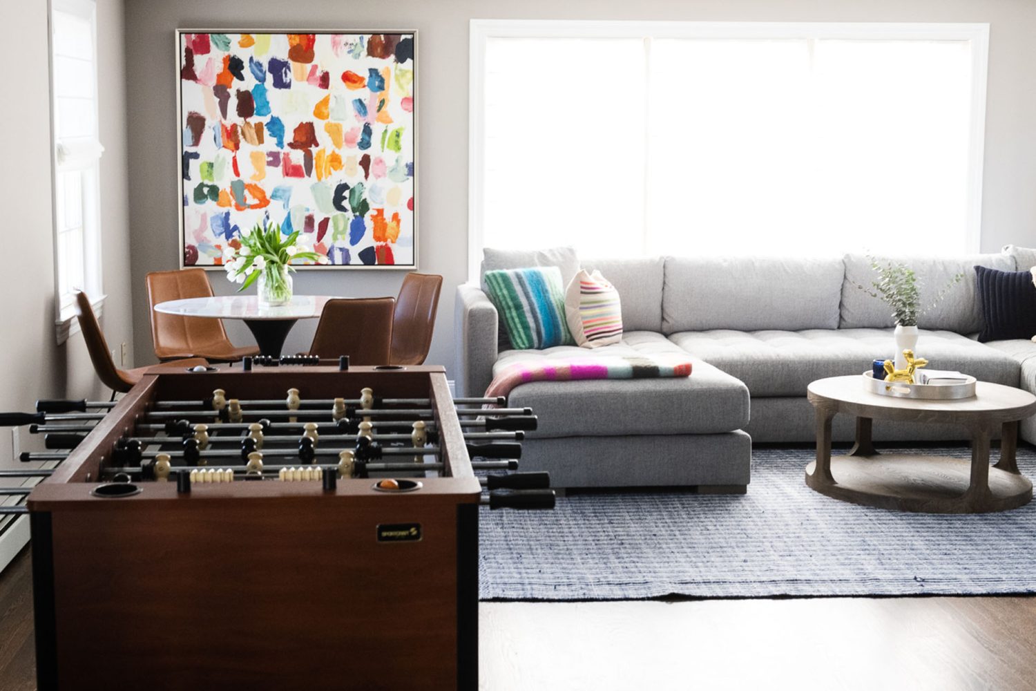 A large, square abstract painting of rainbow-colored brushstrokes above a white game table with leather chairs and aside a light gray sectional couch. There is a medium wood foosball table in the foreground.
