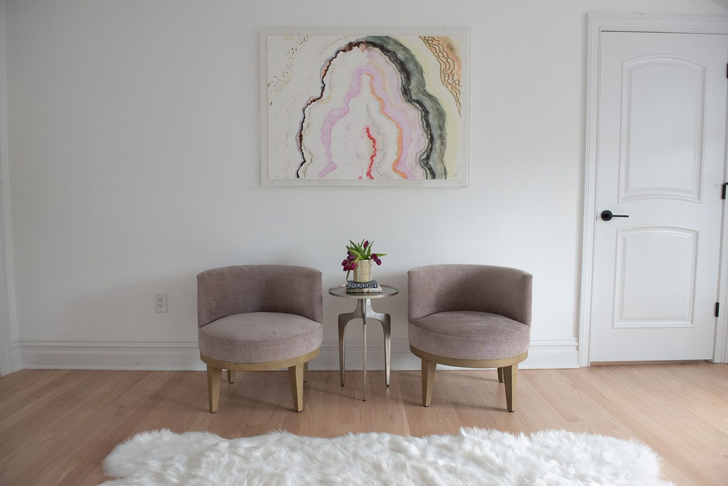 White wall with white deep shag throw rug on light wood floor, pair of taupe upholstered chairs with modern occasional table in between, pastel abstract art on wall.