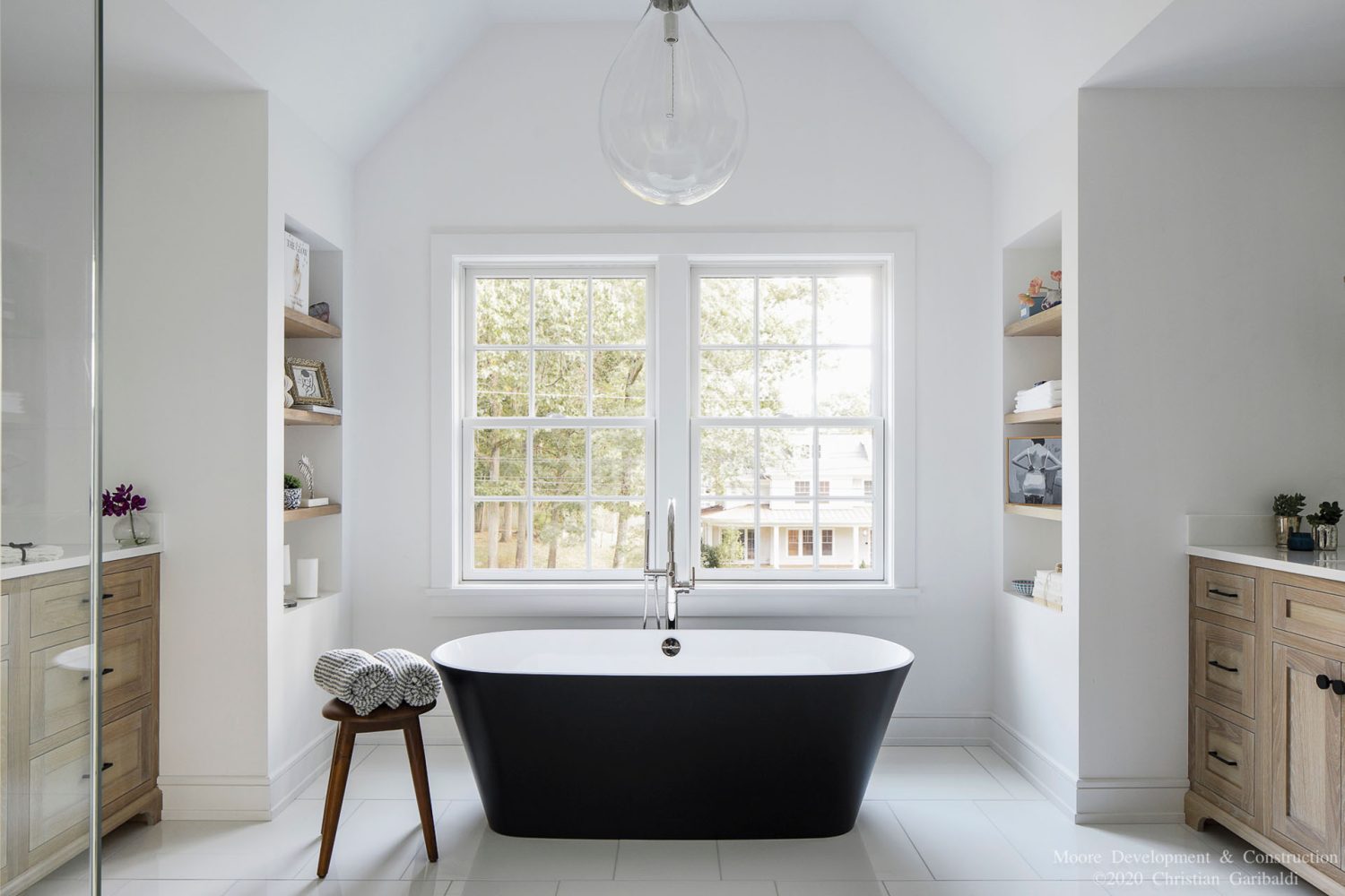 A white-walled bathroom with a modern, freestanding tub that is black outside and white inside. There is a small wooded stool near the tub with two rolled, striped towels on top.
