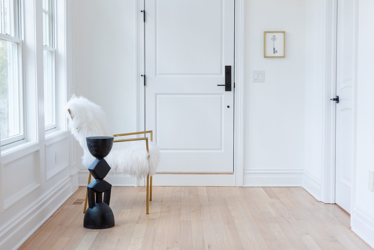A hallway with white walls and light wooden floors. A brass chair with fuzzy white upholstery is set in the corner, next to a sculptural black side-table-height form.