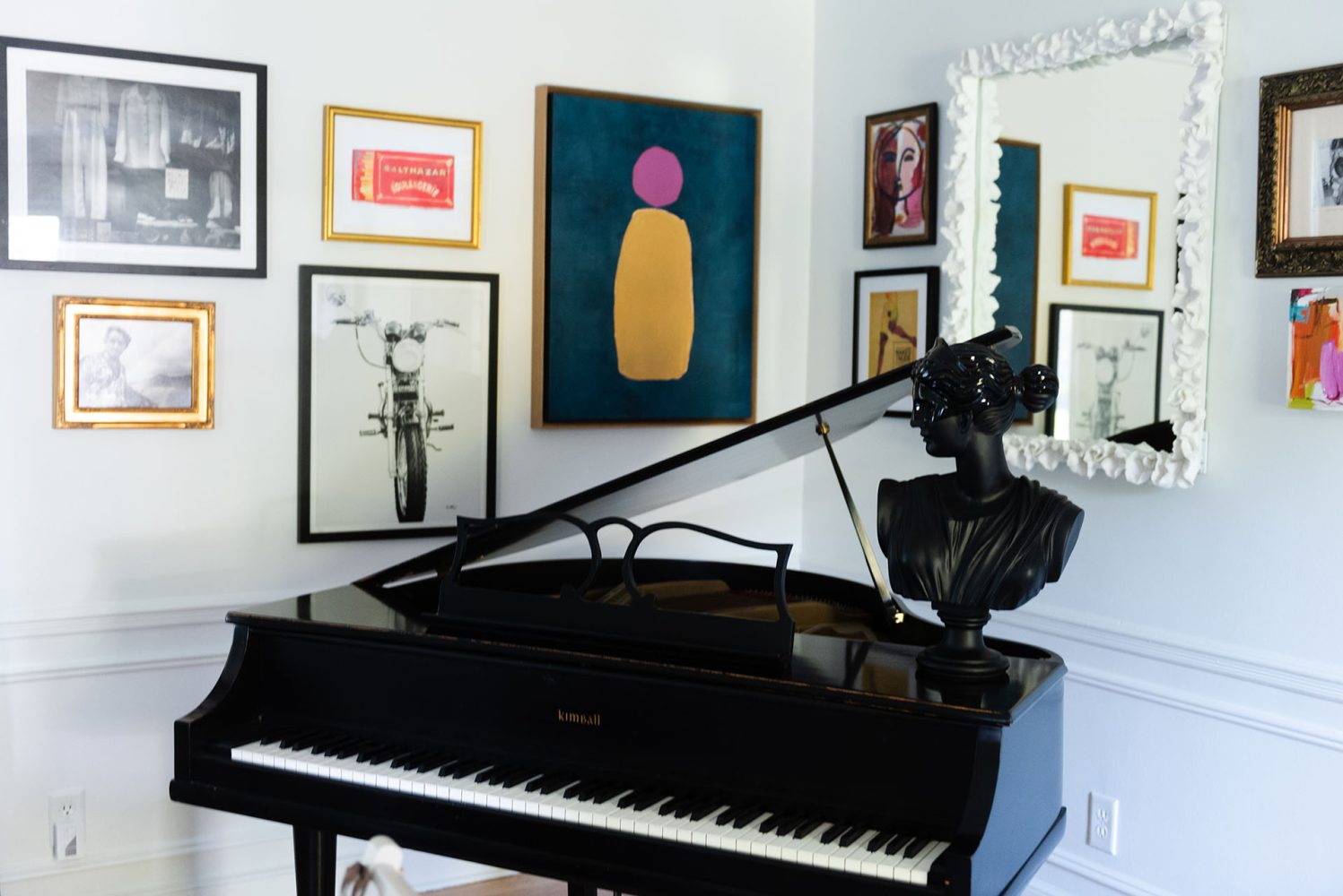 A gallery wall hung in a corner above a black baby grand piano. Art pieces include black and white photography, color paintings and prints, a mirror with a white frame, and a black bust on the piano.