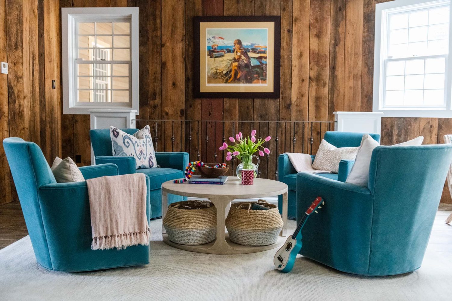 Wood-paneled family room with white-trimmed windows and light area rug. Four medium blue upholstered chairs are arranged around a round coffee table.
