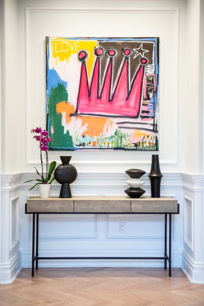 Painting of a pink crown with the word LOVE to the left, against a multicolor background, above a modern beige and steel console decorated with black vases and an orchid.