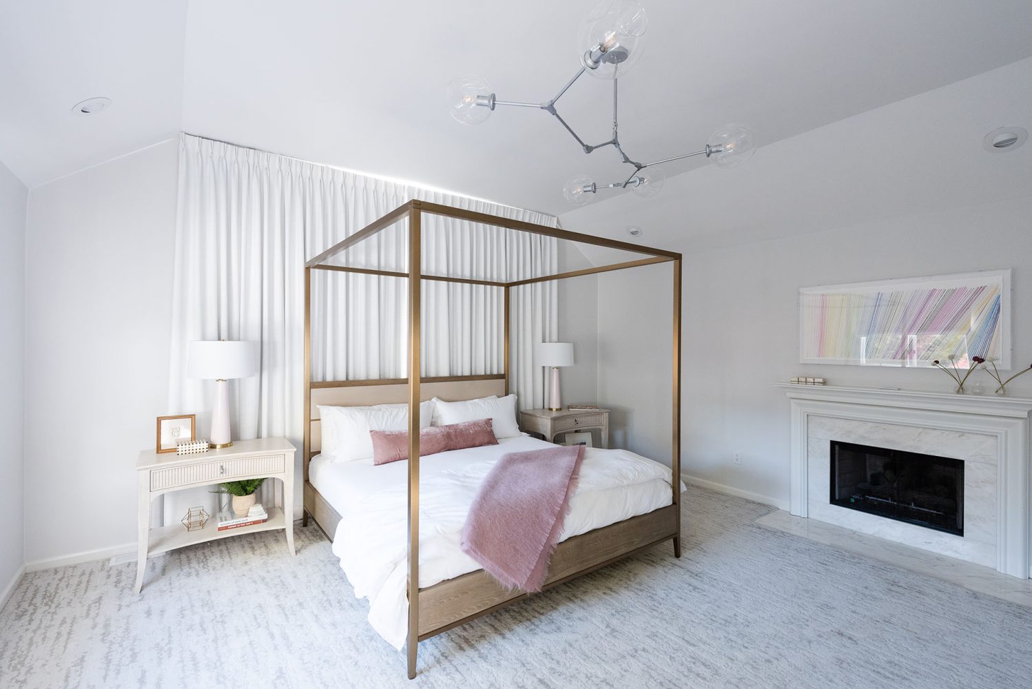 White-walled bedroom with white and grey patterned carpeting. Large, modern brass and wood canopy bed with white bedding and mauve bolster pillow and throw. Modern sliver chandelier, white marble surround fireplace.