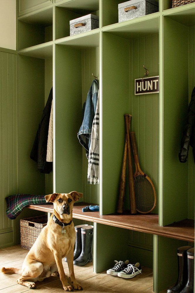 Green locker cubbies in a mudroom. Lower cabinets feature rainboots, sneakers, and a basket. Jackets, a scarf, and a wood sign saying hunt are hung on coathooks. Metal bins are on top shelves.