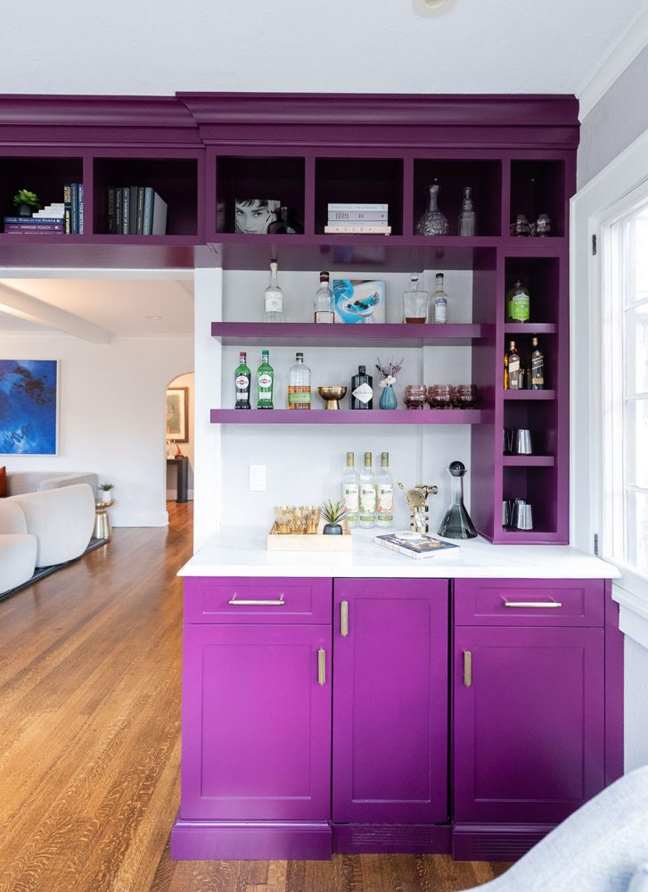 Built-in wood bar, painted purple with brass handles and white countertop. Drawers and cabinets below counter, shelves to the right side and above.