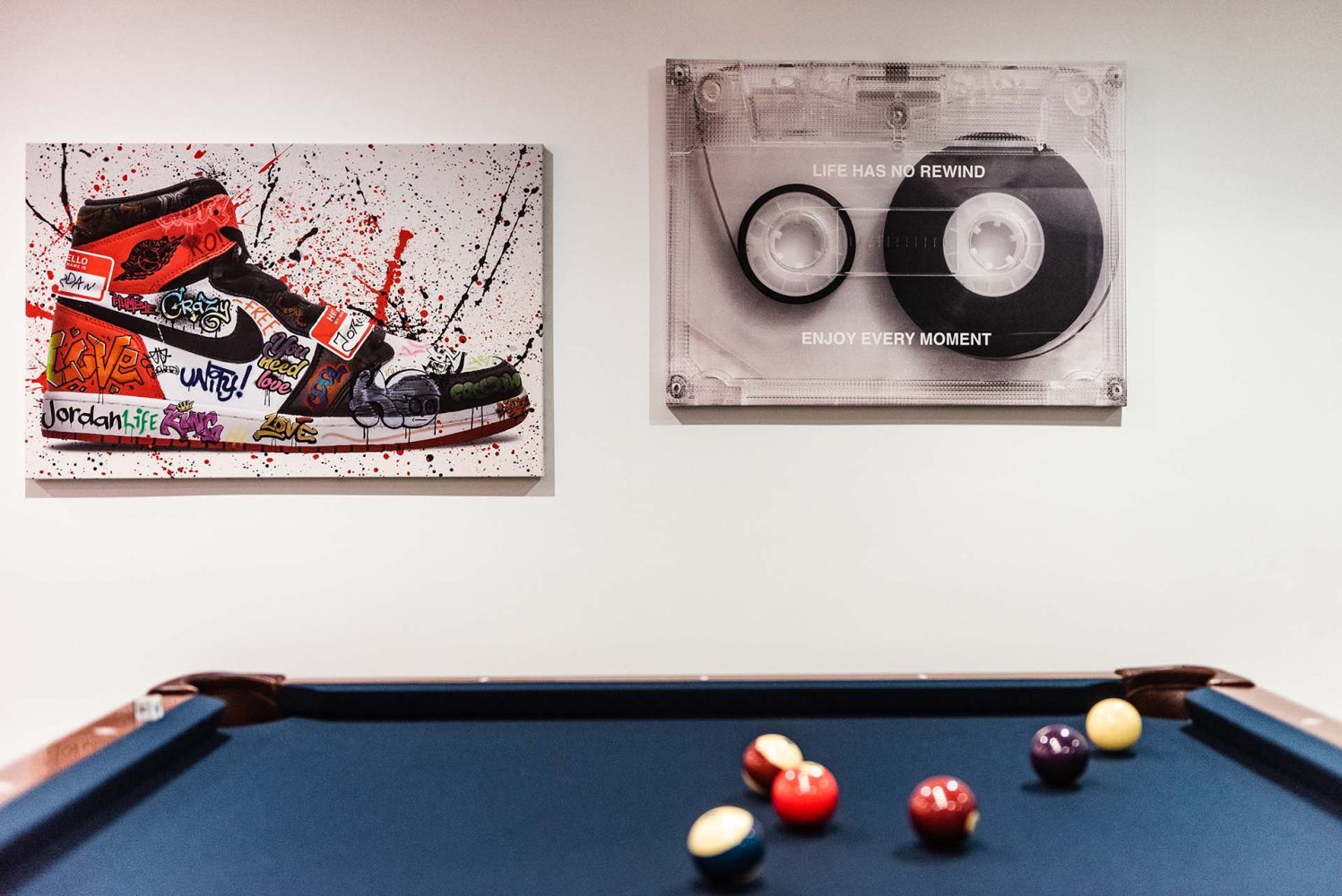 Two art pieces above a pool table. Piece on the left has an Air Jordan sneaker with graffiti and splatter paint, piece on the right is a gigantic clear cassette tape that says Life Has No Rewind Enjoy Every Moment.