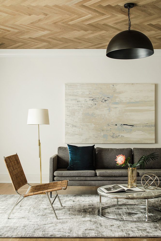 Room with light wood ceiling in a chevron pattern, tan woven leather side chair, chrome and marble round coffee table, grey textured modern sofa, and abstract art in light tan, cream and blues.