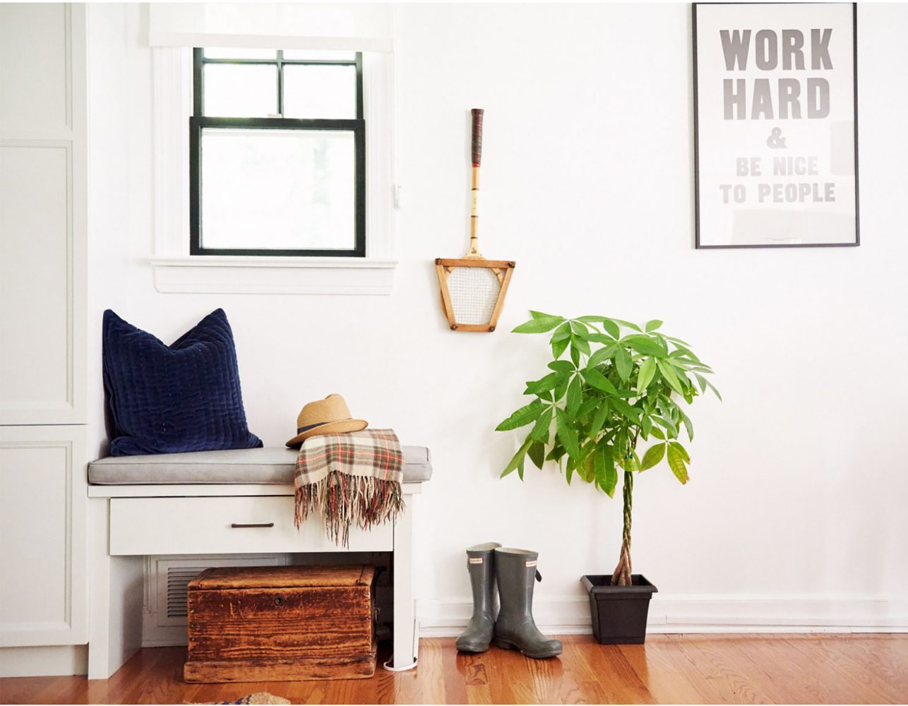 A nook in a room with white walls and medium wood floors. There is a small bench with a gray cushion, navy blue pillow, plaid throw blanket. A vintage tennis racquet hangs on the wall next to a print that says Work Hard & Be Nice To People.